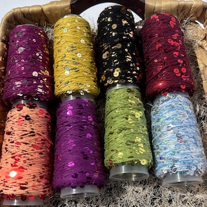 May include: Spools of colourful thread with sequins woven in. The spools are arranged in a basket. The colours are red, orange, purple, green, blue, black, and yellow.