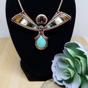 May include: A copper wire wrapped necklace with a turquoise stone pendant and a brown and black stone accent. The necklace is in the shape of a bird with wings spread out.