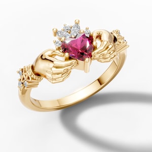May include: A gold-colored Claddagh ring featuring a heart-shaped, deep pink gemstone. The heart is topped with a crown and held by two hands. Small, clear gemstones are set around the crown and on the ring's shoulders.