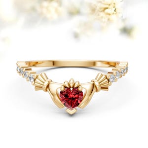 May include: A gold Claddagh ring with a heart-shaped, deep red gemstone at the centre. The ring features small, clear gemstones along the band and stylised hands holding the heart. The ring is set against a soft, blurred background.