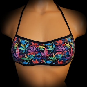 May include: A halter-style crop top with a black trim and straps. The top features a vibrant pattern of colorful cannabis leaves in shades of green, purple, red, and blue against a dark background. The design is eye-catching and bold.