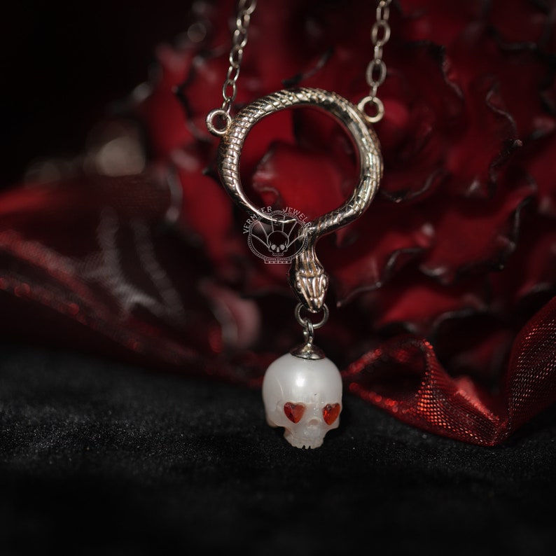 May include: A silver snake necklace with a white skull pendant. The skull has red heart-shaped eyes.