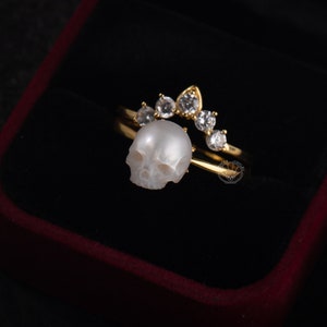 Op de afbeelding: Een gouden ring met een witte parel schedel en een rij heldere zirkonia stenen.