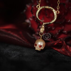 May include: A gold snake necklace with a pearl skull pendant. The skull has red heart-shaped eyes.
