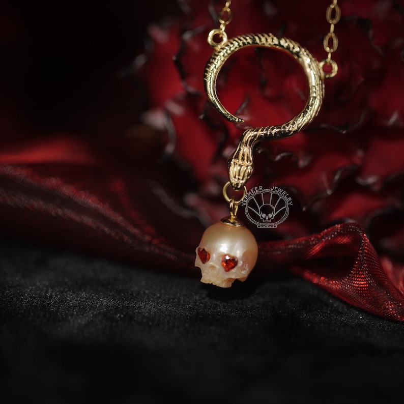 May include: A gold snake necklace with a pearl skull pendant. The skull has red heart-shaped eyes.