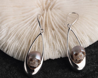 Puede incluir: Un par de pendientes de plata con un dije de calavera nacarada que cuelga de cada pendiente.