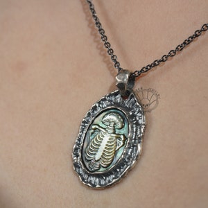 Op de afbeelding: Een zilveren ketting met een hanger met een gedetailleerd skeletontwerp van een zeedier in een getextureerd ovaal frame. De hanger is gemaakt van abalone-schelp met iriserende kleuren.