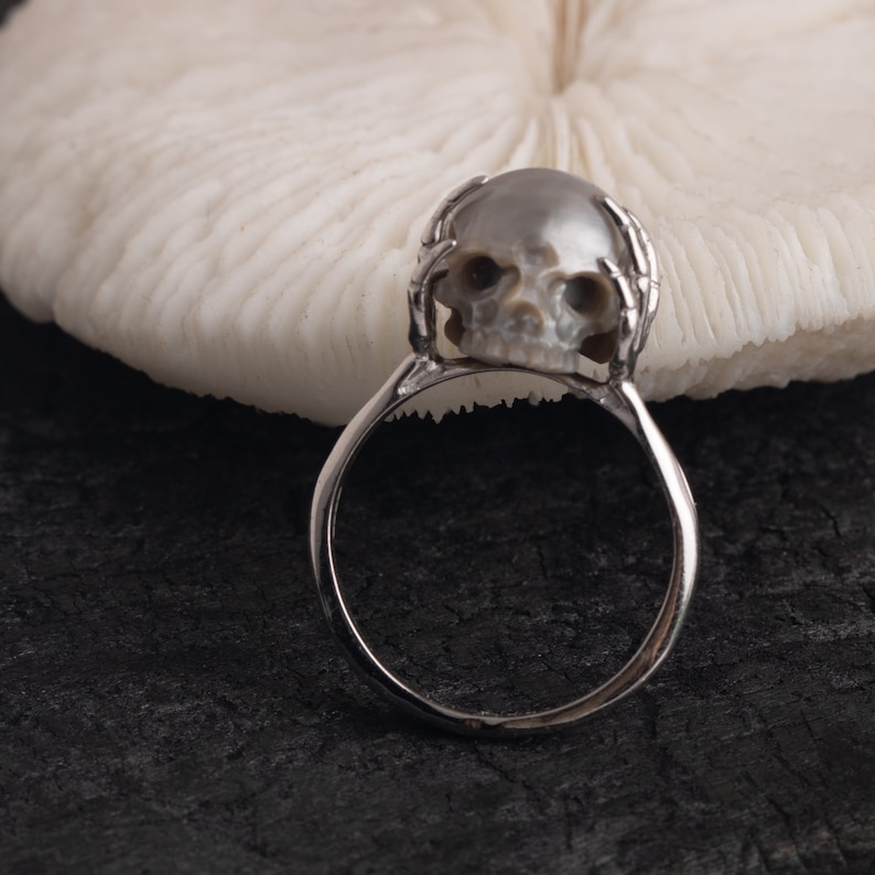 May include: A silver ring with a pearl skull design. The skull is detailed with eye sockets and a nose. The ring is on a white coral background.