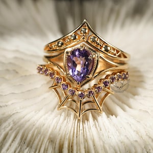 May include: A gold ring set with a purple gemstone, featuring a delicate spiderweb design and a crown-like band with skull details.