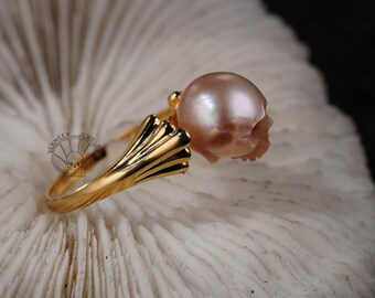 May include: A gold ring featuring a skull design and a large pink pearl. The ring is displayed on a white coral background.