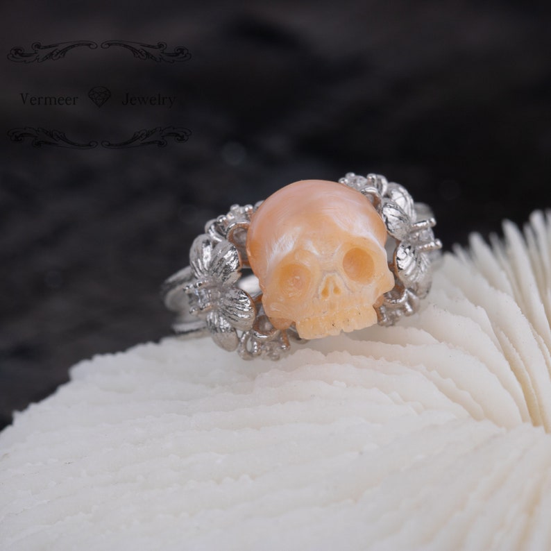 May include: A silver ring with a carved skull made of pink mother of pearl. The skull is surrounded by delicate floral designs.
