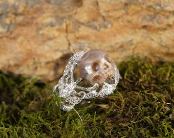 Puede incluir: Un anillo de plata con un diseño detallado que presenta una calavera hecha de nácar iridiscente. El anillo está colocado sobre un fondo de musgo verde.