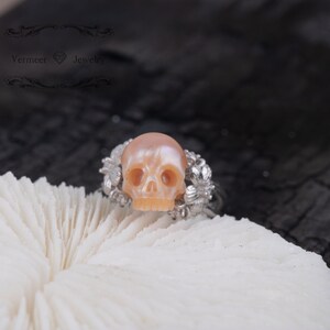 May include: A skull ring made of pink mother of pearl with silver floral accents. The ring is set on a white coral background.