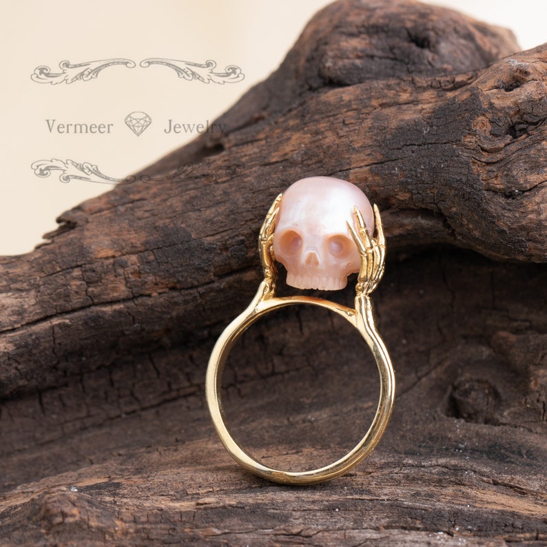 May include: A gold ring with a pink skull made of pearl and two skeletal hands holding the skull.