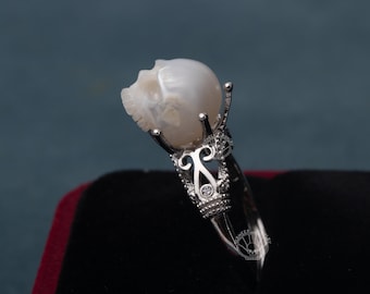May include: A silver ring featuring a carved skull made of a light-colored stone. The skull is held by ornate silver prongs and a decorative band. The ring is set against a dark background.