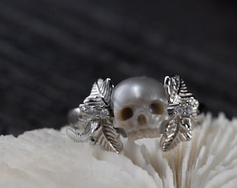 Puede incluir: Un anillo de plata con un cráneo de perla y detalles de hojas. El anillo está sobre un fondo de coral blanco.