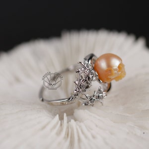 Può includere: Un anello d'argento con un teschio di perla e un designo floreale. La perla è di colore arancio chiaro e i fiori sono decorati con piccole pietre trasparenti.