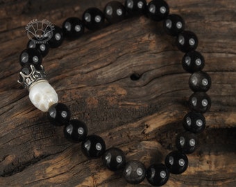 Puede incluir: Pulsera de cuentas negras con un dije de calavera de plata con una corona en la parte superior. La pulsera está sobre una superficie de madera.