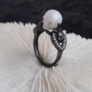 Può includere: Un anello a forma di teschio nero con una perla bianca in cima. L'anello è decorato con piccole pietre chiare.