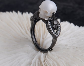 Puede incluir: Un anillo de calavera negro con una perla blanca en la parte superior. El anillo está adornado con pequeñas piedras transparentes.