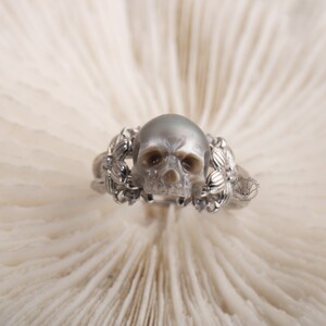 May include: A silver ring with a pearl skull and floral accents. The ring is set on a white background.