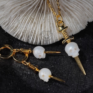 K&ouml;nnte beinhalten: Goldfarbenes Schmuckset mit einer Totenkopf-Anh&auml;nger-Halskette und passenden Ohrringen. Die Totenkopf-Anh&auml;nger sind wei&szlig; und detailliert, mit einem goldfarbenen Dolch-Design. Die Ohrringe haben einen Klappverschluss. Das Schmuckst&uuml;ck wird auf dunklem Untergrund pr&auml;sentiert.