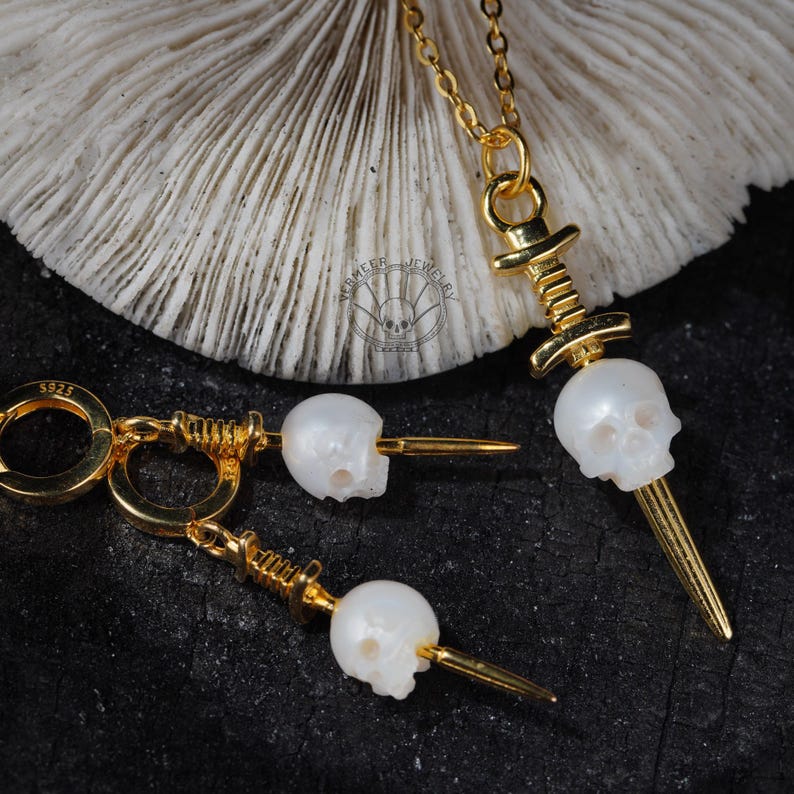K&ouml;nnte beinhalten: Goldfarbenes Schmuckset mit einer Totenkopf-Anh&auml;nger-Halskette und passenden Ohrringen. Die Totenkopf-Anh&auml;nger sind wei&szlig; und detailliert, mit einem goldfarbenen Dolch-Design. Die Ohrringe haben einen Klappverschluss. Das Schmuckst&uuml;ck wird auf dunklem Untergrund pr&auml;sentiert.