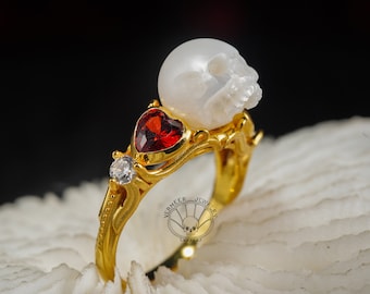 May include: A gold ring featuring a white skull, a red heart-shaped gemstone, and a smaller clear gemstone. The ring is displayed on a white coral-like surface.