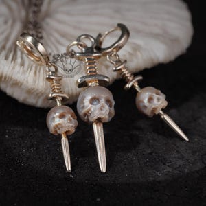 K&ouml;nnte beinhalten: Goldfarbene Sch&auml;del-Ohrringe und eine Anh&auml;nger-Halskette. Die Ohrringe haben kleine Sch&auml;delperlen mit einem Dolch-Design. Die Halskette hat einen &auml;hnlichen Sch&auml;delanh&auml;nger. Der Schmuck wird vor einem wei&szlig;en Hintergrund pr&auml;sentiert.