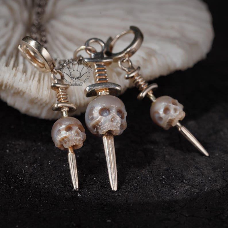 K&ouml;nnte beinhalten: Goldfarbene Sch&auml;del-Ohrringe und eine Anh&auml;nger-Halskette. Die Ohrringe haben kleine Sch&auml;delperlen mit einem Dolch-Design. Die Halskette hat einen &auml;hnlichen Sch&auml;delanh&auml;nger. Der Schmuck wird vor einem wei&szlig;en Hintergrund pr&auml;sentiert.