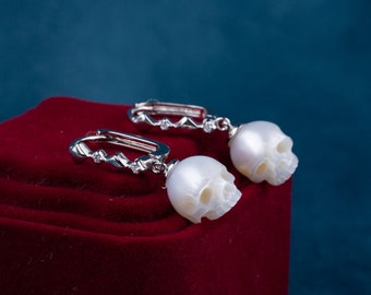 Puede incluir: Un par de pendientes de plata con forma de calavera con detalles de perlas blancas. Los pendientes están sobre un fondo de terciopelo rojo.