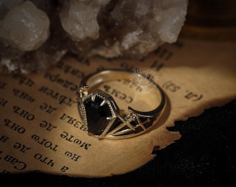 May include: A silver ring with a black coffin-shaped stone. The ring has black geometric designs on the band. The ring is set on a piece of aged paper with text.