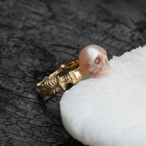 May include: A gold ring featuring a skull design and a pearl skull charm. The ring is displayed on a white surface.