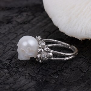 May include: A silver ring with a white pearl skull and a flower design. The ring is on a dark wooden surface.