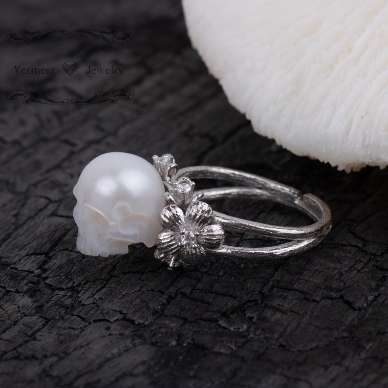 May include: A silver ring with a white pearl skull and a flower design. The ring is on a dark wooden surface.