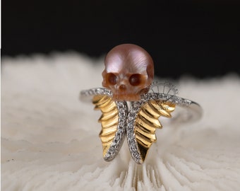 Puede incluir: Un anillo de plata con una calavera de perla y alas doradas engastadas con diamantes. El anillo está sobre un fondo de coral blanco.