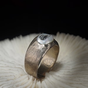 May include: A silver ring with a textured band and a white shell inlay. The ring is set on a white coral surface. The text "VERMEER JEWELRY" is engraved on the band.