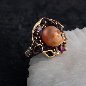 Peut inclure: Une bague en or avec un crâne sculpté en coquille orange. Le crâne est entouré d'un cadre en métal noir avec de petites pierres précieuses roses.