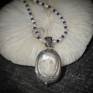 May include: A silver locket necklace with a white mother of pearl skull design. The necklace is made of white pearls and blue beads.