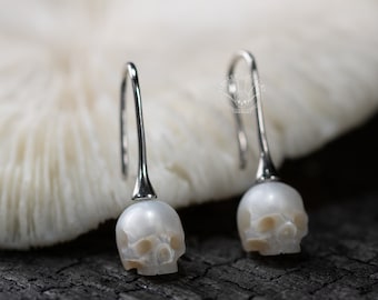 May include: A pair of silver dangle earrings with white pearl skull charms. The earrings are hanging from a white coral-like surface.