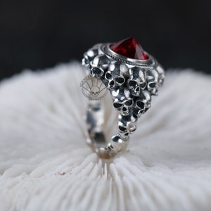 Puede incluir: Un anillo de plata con una piedra preciosa roja rodeada de un grupo de diseños de calaveras.