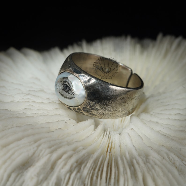 May include: A silver ring featuring a large, oval-shaped, white pearl-like stone set into the top. The ring has a textured, hammered finish and is displayed on a white coral-like surface.