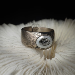 May include: A silver ring with a textured band and a white oval inlay featuring a black eye design. The ring is on a white coral-like surface.