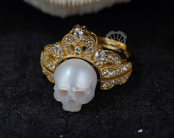 May include: A gold ring with a skull-shaped pearl, set under a crown design with clear gemstones. The ring is displayed on a dark, textured surface, highlighting the intricate details of the jewellery.