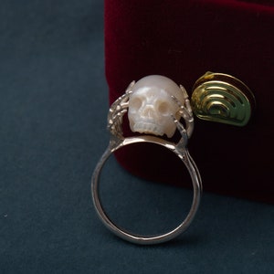 May include: A silver ring with a white pearl skull design. The ring is set against a red velvet background.