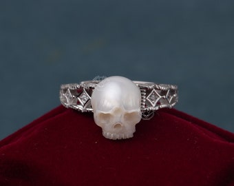 Puede incluir: Un anillo de plata con un diseño de calavera de perla blanca. El anillo tiene una banda delicada e intrincada con acentos de diamantes.
