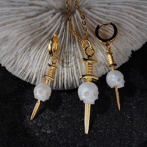 K&ouml;nnte beinhalten: Ein goldfarbenes Schmuckset mit einer Halskette und zwei Ohrringen. Jedes St&uuml;ck hat einen wei&szlig;en Totenkopfanh&auml;nger mit einem goldfarbenen Dolch. Die Ohrringe sind im Creolen-Stil. Die Halskette hat eine zarte Kette.