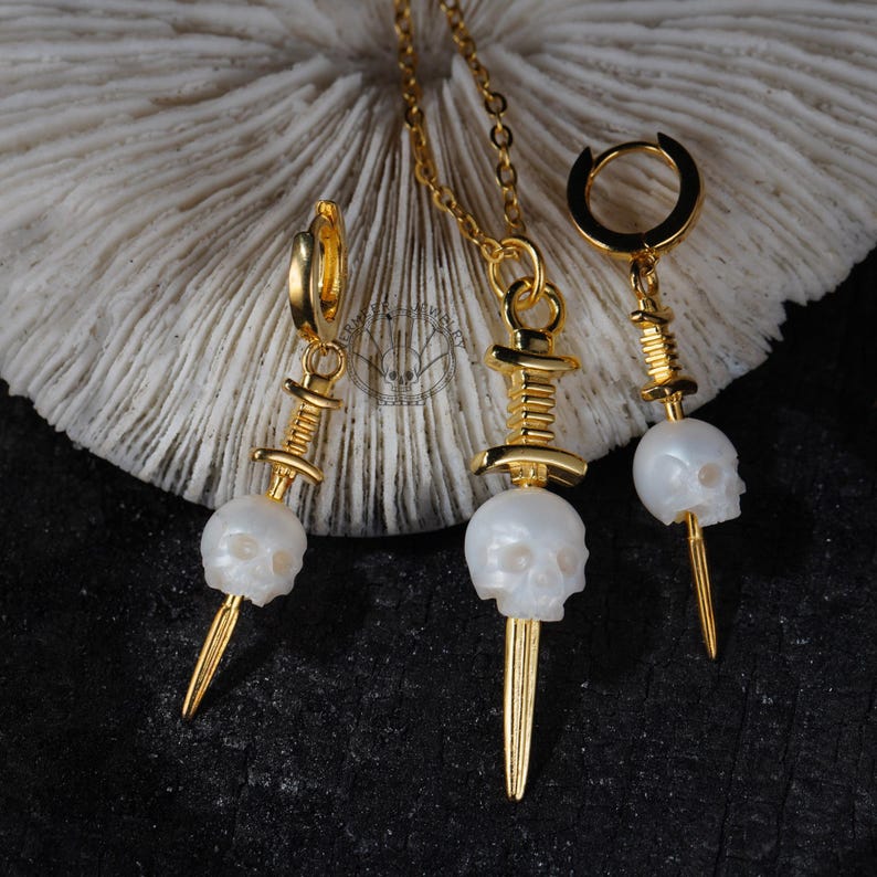 K&ouml;nnte beinhalten: Ein goldfarbenes Schmuckset mit einer Halskette und zwei Ohrringen. Jedes St&uuml;ck hat einen wei&szlig;en Totenkopfanh&auml;nger mit einem goldfarbenen Dolch. Die Ohrringe sind im Creolen-Stil. Die Halskette hat eine zarte Kette.