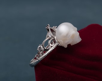 Puede incluir: Un anillo de plata con un diseño de calavera de perla. El anillo está colocado sobre un fondo de terciopelo rojo.