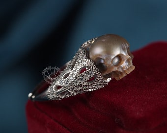 Puede incluir: Un anillo de plata con una perla en forma de calavera. El anillo está engastado con pequeños diamantes y tiene un diseño delicado e intrincado.